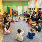 Dzieci i nauczycielka siedzą w kole i trzymają kolorowe chusteczki. Obok dzieci na krzesłach siedzą rodzice. Dookoła znajdują się przedszkolne zabawki.