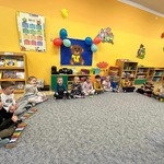 Dzieci w półokręgu siedzą na dywanie i trzymają instrumenty. Za dziećmi znajdują się zabawki oraz dekoracja: miś i balony.
