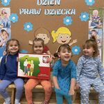 Cztery uśmiechnięte dziewczynki siedzą na ławce. Jedna z nich trzyma planszę z mapą Polski i Godłem Polski. Dzieci są ubrane w kolorze niebieskim. Z tyłu dekoracja z okazji Międzynarodowego Dnia Praw Dziecka.