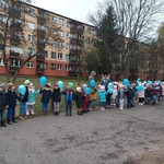 Dzieci stoją w szeregu i trzymają w dłoniach niebieskie balony oraz pompony. Za dziećmi widać blok i drzewa. 