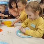 1.	Dziewczynka w żółtej bluzce siedzi przy stoliku, lepi z masy plastycznej pizzę.