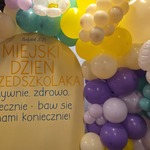 Tablica z napisem: Miejski Dzień Przedszkolaka. Aktywnie, zdrowo, tanecznie – baw się z nami koniecznie. Obok kolorowe balony.