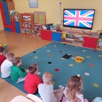 Na zdjęciu widzimy dzieci, które siedzą w rzędzie przed tablicą interaktywną, na której jest wyświetlana flaga Wielkiej Brytanii. 