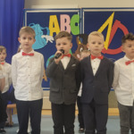 Na zdjęciu widzimy dzieci, które są ubrane w galowe ubrania. Jeden z chłopców trzyma mikrofon. Za dziećmi znajduje się dekoracja z okazji Dnia Edukacji Narodowej.