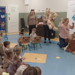 Na środku siedzą na krzesłach dwaj chłopcy i trzymają w rękach sznurek z kartonowym miodem, nawijają go na rulon przez głowę misia. Obok stoją nauczycielki a po obu stronach siedzą dzieci i kibicują im w zawodach.