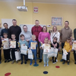 Dzieci stoją z dyplomami i nagrodami za nimi rodzice.