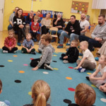 Na dywanie siedzą dzieci, które są odwrócone przodem do nauczycielki. Za dziećmi na krzesłach siedzą rodzice. Dzieci patrzą na nauczycielkę, która klaszcze w dłonie. 