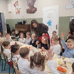 Elegancko ubrane dzieci siedzą przy wigilijnym stole. Nauczycielka trzyma mikrofon przez który dziewczynka odpowiada na zadane pytanie.