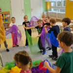 Dzieci stoją w kole i trzymają w rękach kolorowe chustki. Przed nimi stoi prowadząca.