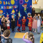 Dzieci tańczą ustawione w kole. Na drugim planie widać karnawałową dekorację.