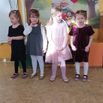 Elegancko ubrane dziewczynki stoją w szeregu, a za nimi widać dekorację z okazji Dnia Babci i Dziadka.