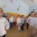 Dzieci ubrane w stroje galowe, stoją ustawione w szachownice. Na drugim planie widać dekorację z okazji Dnia Babci i Dziadka.