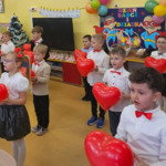Dzieci, które są ubrane w stroje galowe, stoją ustawione w szachownice i trzymają balony w kształcie serc. Za dziećmi widać dekorację z okazji Dnia Babci i Dziadka.