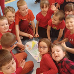 Dzieci ubrane na czerwono siedzą na podłodze, na środku leży pomalowana kartka w kształcie serca, obok widać kilka stojących dzieci.