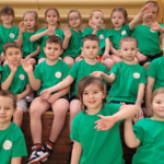 Na trybunach szkolnych siedzi grupa przedszkolaków ubrana w krótkie spodenki i zielone koszulki z logo przedszkola. Dzieci machają ręką.