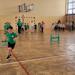 Zdjęcie przedstawia chłopca, który biegnie pokonując przeszkody- płotki gimnastyczne. W tle widzimy ustawione drużyny gotowe do zawodów sportowych.