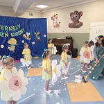 Na scenie stoją dziewczynki ubrane w żółte koszulki i żółto-zielone spódniczki. W dłoniach trzymają duże papierowe kwiaty. Przed nimi nauczycielka w zielonej kwiecistej sukience i wianku na głowie zamiata białe papierowe śnieżynki miotłą oklejoną kolorowymi kwiatami. W tle widzimy dekorację wiosenną.