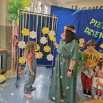 Na zdjęciu widzimy nauczycielkę przebraną za Panią Wiosnę. Przed nią stoi dziewczynka i rozwiązuje zagadkę zawieszoną na kolorowym kwiatku. W tle dekoracja wiosenna i inne dzieci biorące udział w quizie.