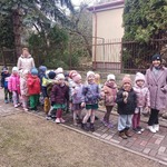 Na zdjęciu dwie panie i grupa dzieci ubranych w kurtki stoją na podwórku. W dłoniach trzymają instrumenty muzyczne i uśmiechają się do aparatu.