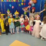 W szeregu stoją uśmiechnięte dzieci w kolorowych strojach: ScoobyDoo, Pikachu, księżniczka, dzwoneczek. Za dziećmi widać radosną kobietę w czerwonym kapeluszu z podniesionymi rękoma. W tle kolorowa dekoracja. 