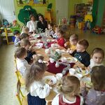 Nauczycielki i dzieci siedzą przy wigilijnym przedszkolnym stole, na którym są świąteczne potrawy i odświętna dekoracja. Nauczycielki i przedszkolaki są ubrane w galowe stroje.