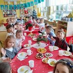 Dzieci siedzą przy wigilijnym przedszkolnym stole, na którym są świąteczne potrawy i odświętna dekoracja. Przedszkolaki są ubrane w galowe stroje.