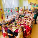 Nauczycielki i dzieci siedzą przy wigilijnym przedszkolnym stole, na którym są świąteczne potrawy i odświętna dekoracja. Nauczycielki i przedszkolaki są ubrane w galowe stroje.