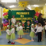 Dzieci odświętnie ubrane tańczą w parach wspólnie z trzema paniami. W tle dekoracja – żółty napis na zielonym tle, kolorowe postacie, balony. 