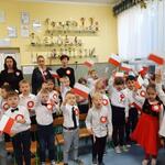 Dzieci stoją ubrane w stroje galowe z przypiętymi kotylionami. Machają flagami Polski. Za dziećmi cztery panie. Wszyscy śpiewają Mazurek Dąbrowskiego.