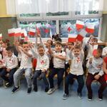 Na krzesełkach siedzą dzieci ubrane w stroje galowe, z przypiętymi kotylionami. Machają flagami Polski.