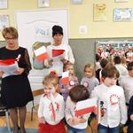 Dzieci stoją ubrane w stroje galowe z przypiętymi kotylionami. Machają flagami Polski. Obok dzieci dwie panie. Wszyscy śpiewają Mazurek Dąbrowskiego.
