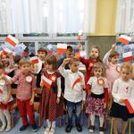 Dzieci stoją ubrane w stroje galowe, z przypiętymi kotylionami. Machają flagami Polski.