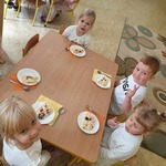 Pięcioro dzieci ubranych na biało siedzi przy stoliku. Przed nimi na stole są talerze z tortem stojące na kolorowych serwetkach.
