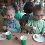 Dzieci ubrane na zielono siedzą przy stolikach i jedzą tort z okazji Dnia Przedszkolaka.