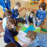 Na dywanie siedzi siedmioro dzieci ubranych na niebiesko. Na środku leży duży arkusz białego papieru. Dzieci wyklejają na arkuszu rzekę i trawę kawałkami niebieskiego i zielonego papieru. W tle niebieskie pudełka z zabawkami. 