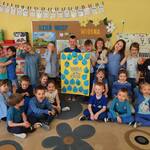 Na dywanie siedzi grupa uśmiechniętych dzieci ubranych na niebiesko. Za nimi stoją dzieci trzymające plakat na żółtym papierze z niebieskimi kroplami wody i napisem „Woda to życie”. W tle dekoracja z okazji Dnia Wody. 