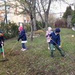 Z przodu dwie dziewczynki i dwóch chłopców grabią trawę. W tle znajdują się skalniaki, drzewa, krzewy, budynek szkoły i blok osiedlowy